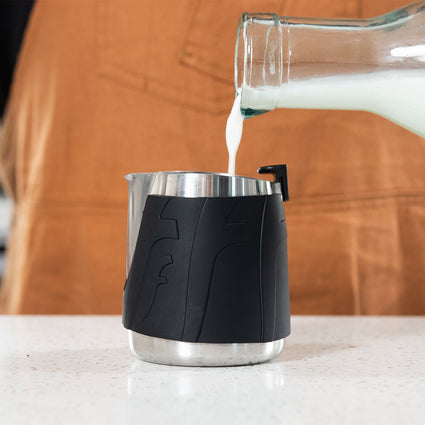 Glass container pouring milk into a black and silver pitcher on a wooden surface.
